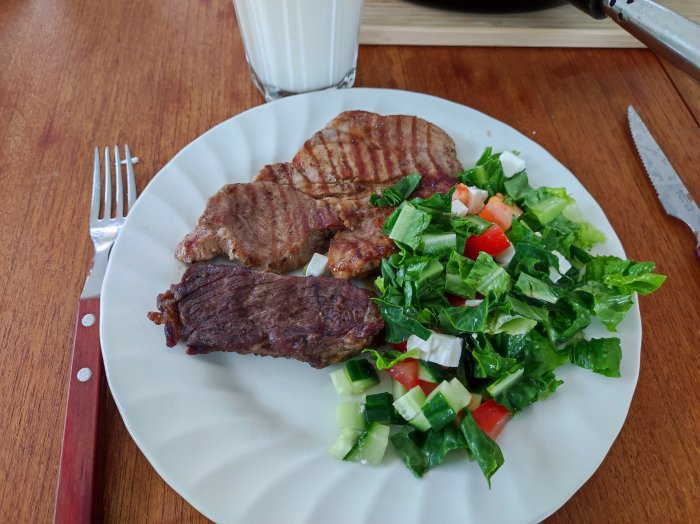 Grillade biffar med färsk grönsallad på en vit tallrik, intill bestick och ett glas mjölk.