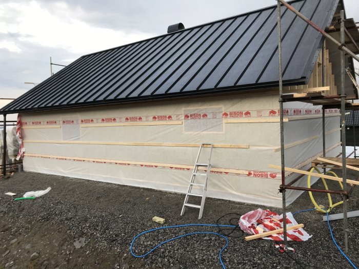 Hus under renovering med isolering och vindduk delvis påsatt, byggmaterial och stegar synliga.