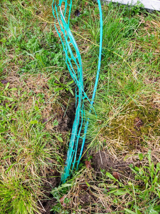 Märkband med metalltråd ovanpå nedgrävd kabel i grävd ränna på gräsmatta.