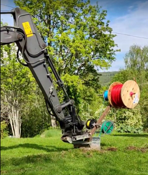 Grävmaskin med pågående fiberkabelinstallation, spolar med kablar och varningsband i trädgård.