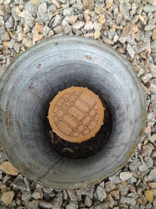 Inspektionsbrunn med brunt lock omgiven av grus, synligt vatten inuti röret.