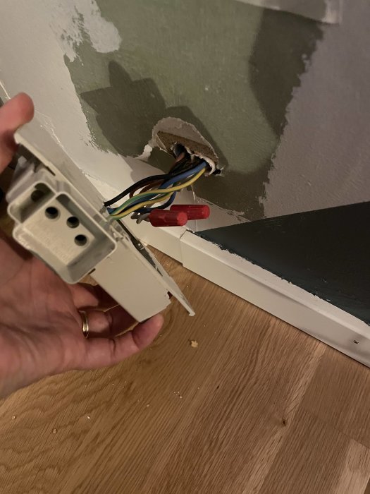 Hand håller en strömbrytare framför ett öppet hål i väggen där elektriska kablar syns.
