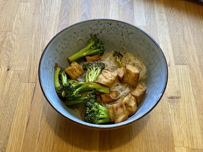 En skål med glasnudlar, stekt broccoli och gyllene tofu på ett trägolv.