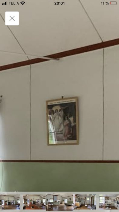 Interiör av missionshus med vita väggpaneler och bruna lister, skarvar och inramad bild synlig.