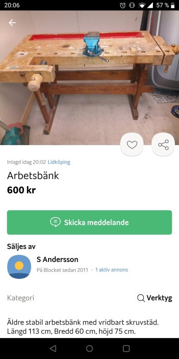 Använd arbetsbänk i trä med blå skruvstycke, synliga slitage och verktyg på golvet.