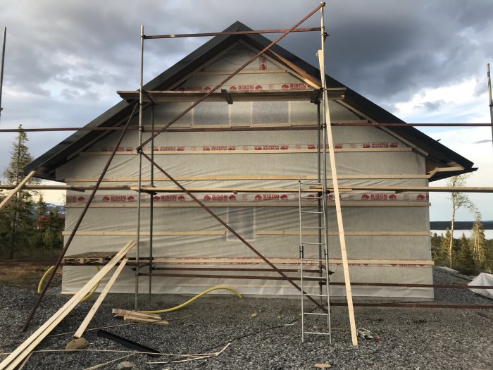 Hus i byggfas väderskyddat med vindduk, ställningar runt och byggmaterial på marken.