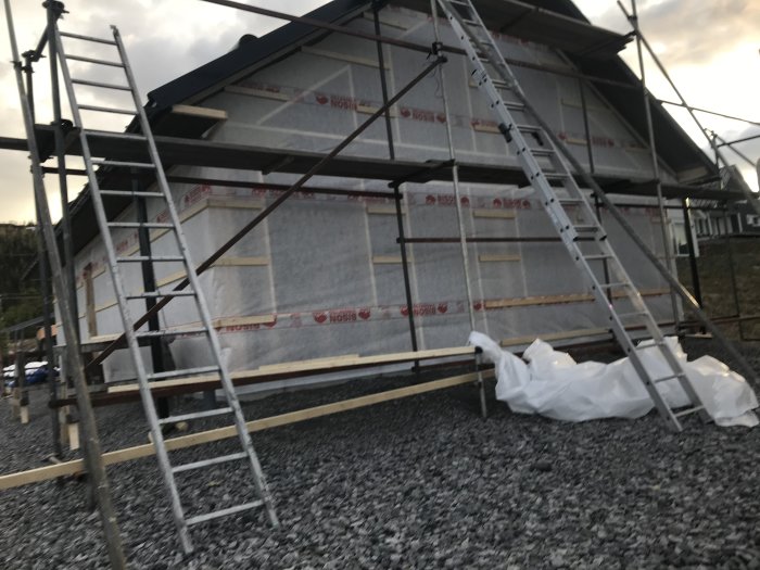 Ett hus under konstruktion med väderskydd bestående av vindduk och spikläkt, omgivet av byggnadsställningar.