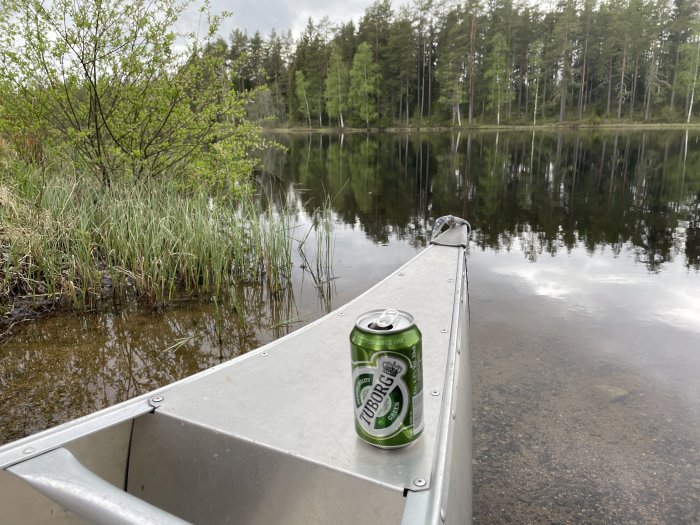 En aluminiumbåt med en ölburk på kanten, med skog och spegelblank sjö i bakgrunden.