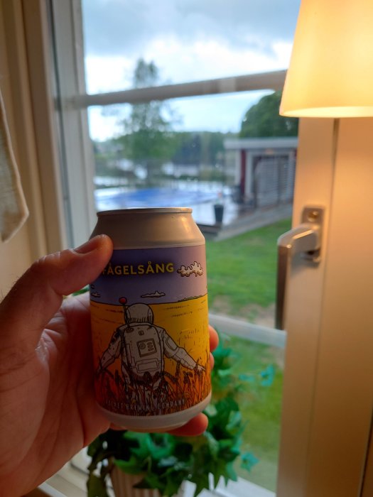 Hand håller en ölburk med konstnärlig design framför fönster med regn ute.