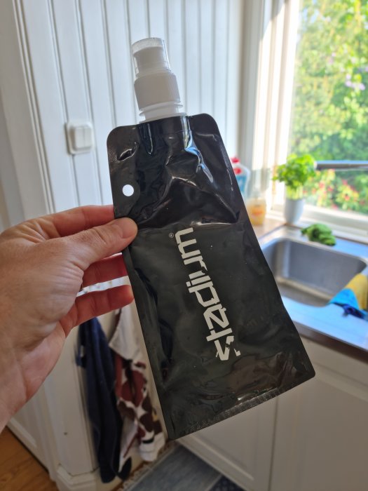 Hand håller en skrynklig svart vattenflaska med vit kork och logotyp som ser läckande ut.