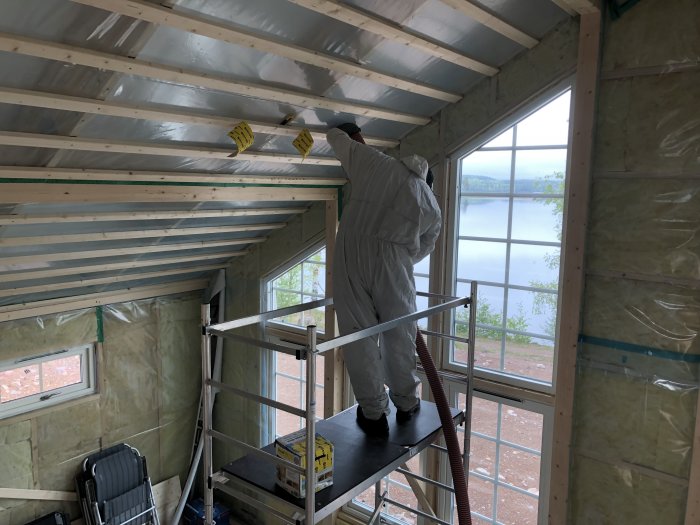 Person i skyddsdräkt blåser isolering i taket på ett hus under renovering med sjöutsikt.