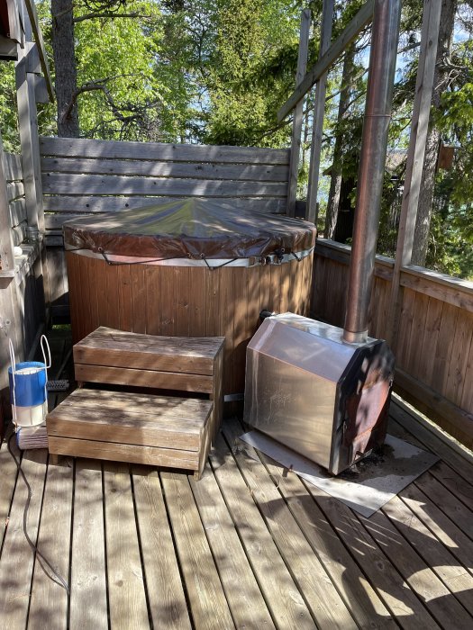 En badtunna med täckt lock på en träterrass, med trappsteg framför och en aluminiumkamin bredvid.