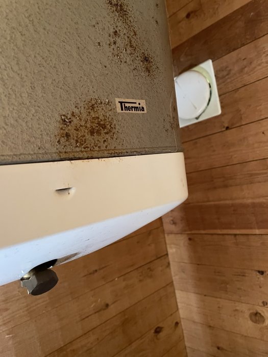 En beige Thermia varmvattenberedare från 70-talet med rostfläckar, placerad på ett trägolv.