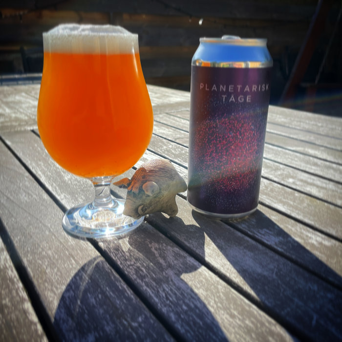 En ölglas fylld med orangefärgad dryck bredvid en burk öl och en små skulptur av en igelkott på ett träbord.