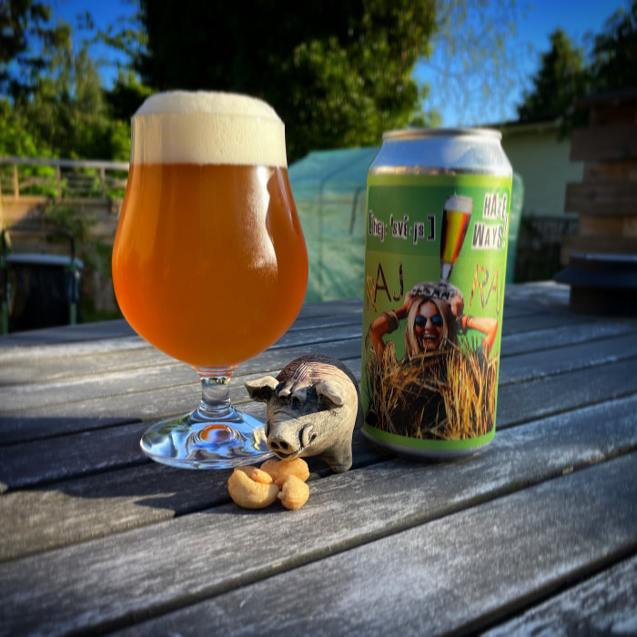 Ölglas med skummad dryck bredvid en ölburk och en trägrisfigur med cashewnötter på ett bord utomhus.