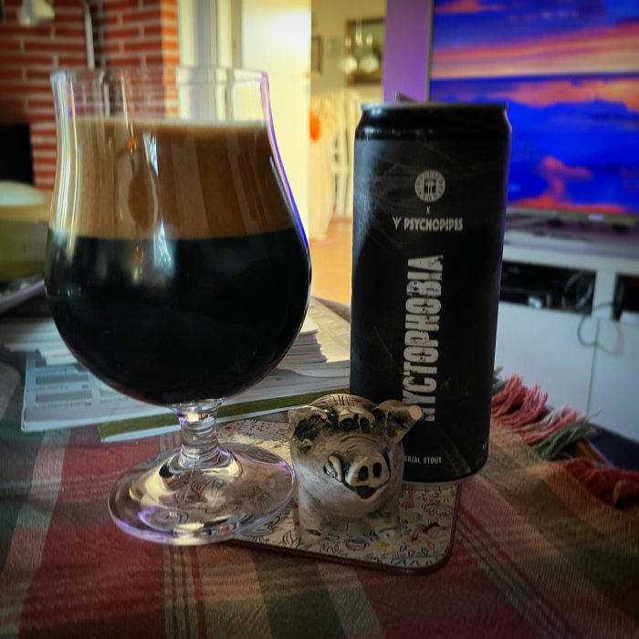 Ett glas mörkt öl med tjock skumkrona framför en burk med texten "PSYCHOP/PS" på ett bord.