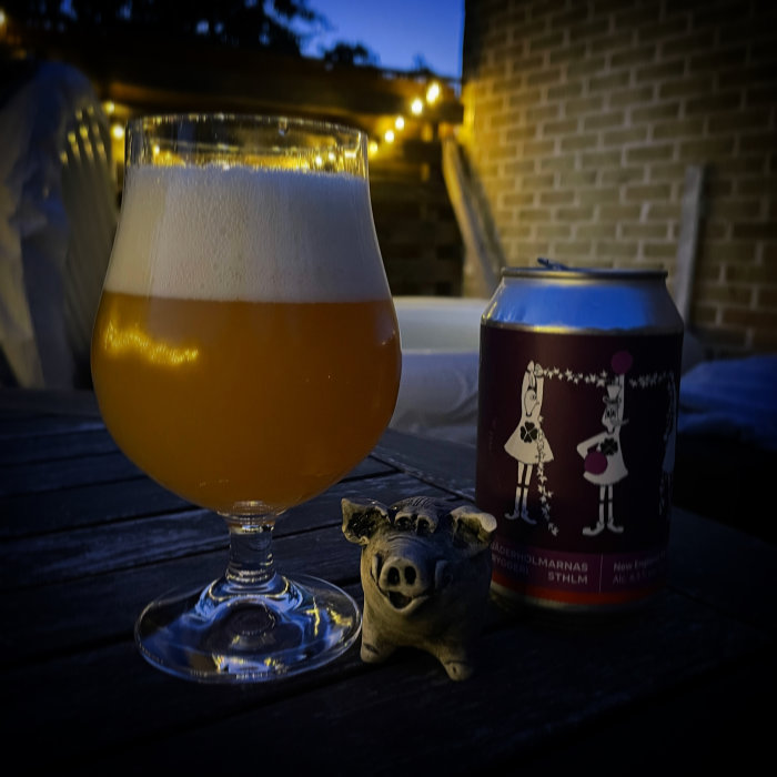 Ett glas öl bredvid en ölburk och en liten prydnadsgris på ett träbord utomhus i skymningen.