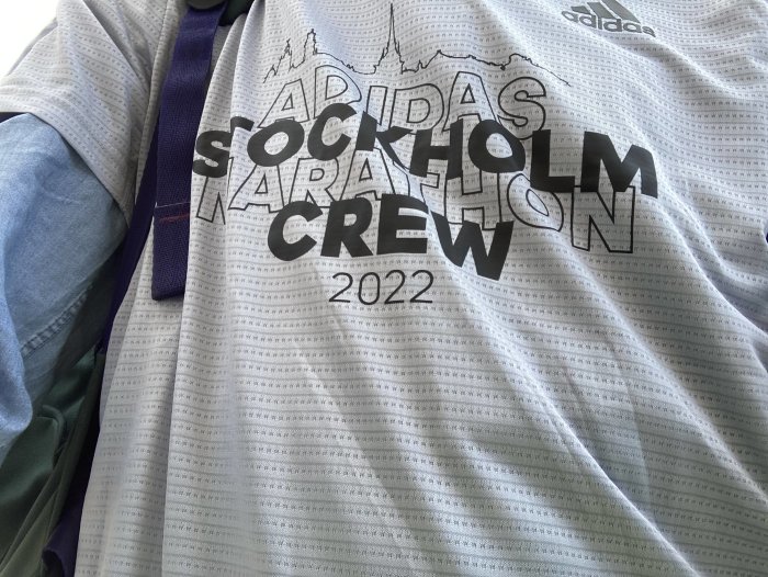 Gråvit Adidas-tröja med texten "ADIDAS STOCKHOLM CREW 2022" framför suddig bakgrund.