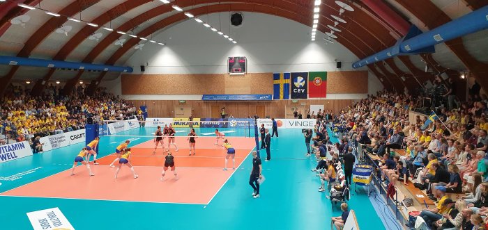 Publik som tittar på en inomhusvolleybollmatch mellan två lag i en sportarena.