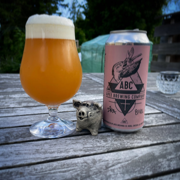 Ölglas med skum topp bredvid ölburk från Apex Brewing och en liten grisskulptur på träbord.