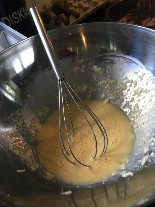 En skål med en visp och en rörd äggsmet till traditionell pasta carbonara, med synliga klumpar av riven ost.