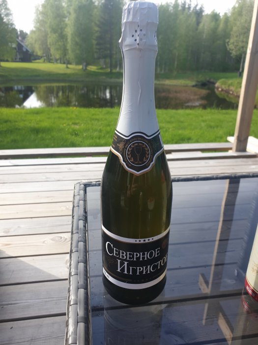 Flaska med rysk "champagne" på ett utedäck med damm och skog i bakgrunden.