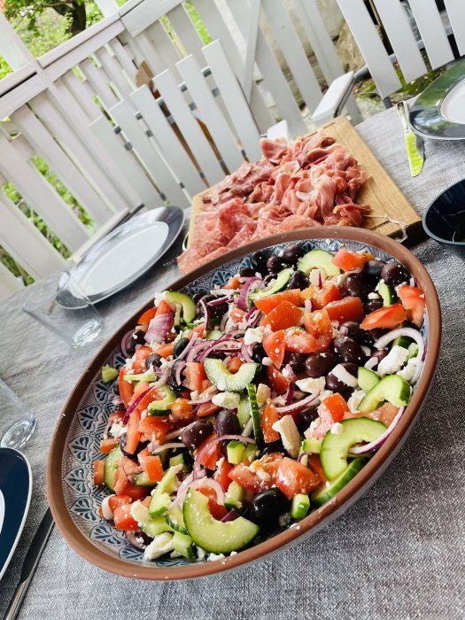 Grekisk sallad med tomat, gurka, oliver och fetaost samt träbricka med charkuterier på ett bord utomhus.