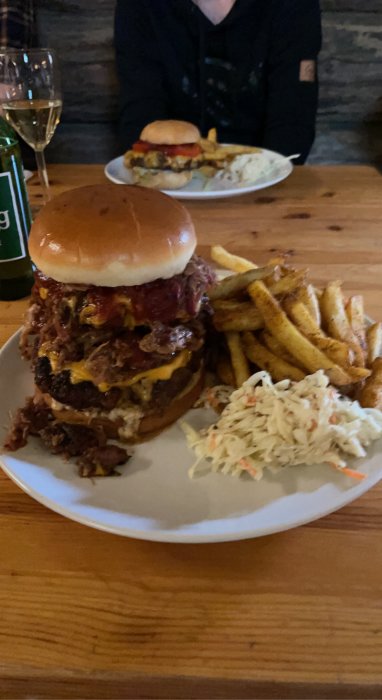 En stor burgare med pulled pork, ost och tillbehör, serverad med pommes och coleslaw på en vit tallrik.