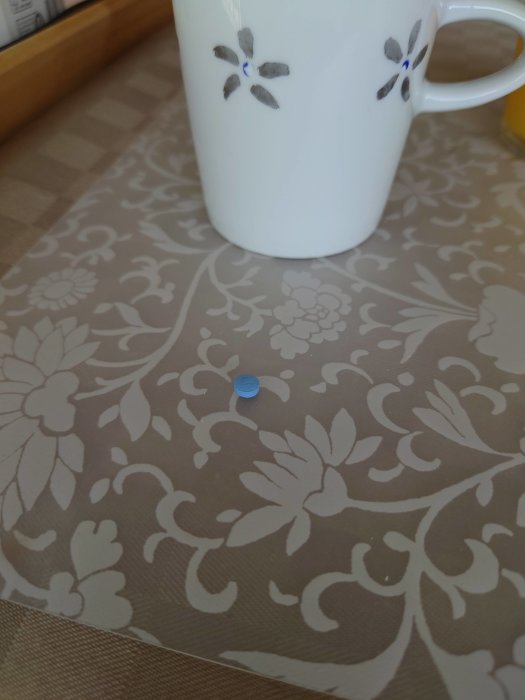 Vit kaffekopp med blomdekoration på ett blommigt bordsunderlägg med en liten blå plastbit.