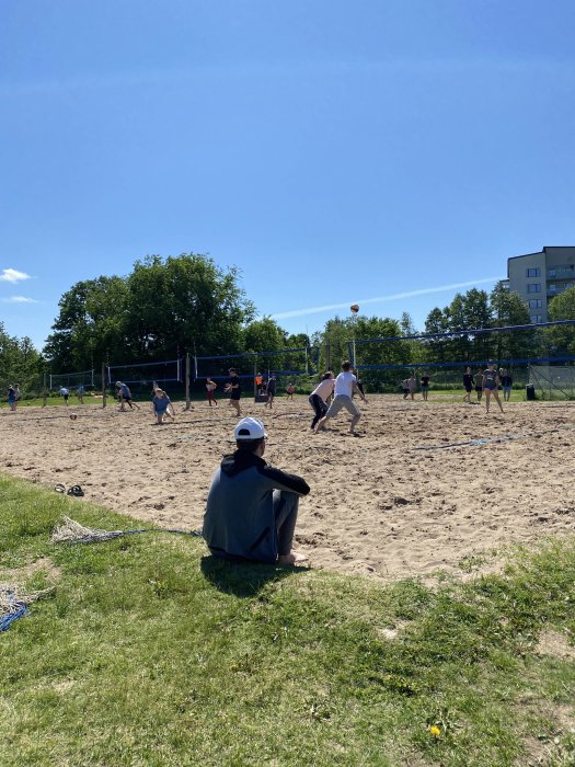 Person i förgrunden tittar på beachvolleybollspelare på en sandplan en solig dag.