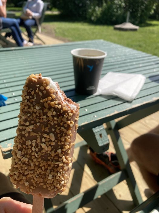 Halväten glasspinne med nötter framför ett kaffemugg på ett utomhusbord i solljus.