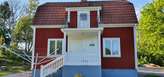 Rött svenskt hus med vita knutar och balkong, omgivet av gröna träd.