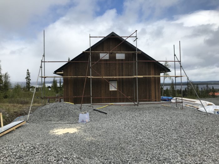 Hus under konstruktion med färdigställd gavel och ställningar, omgiven av grus och skog med sjö i bakgrunden.