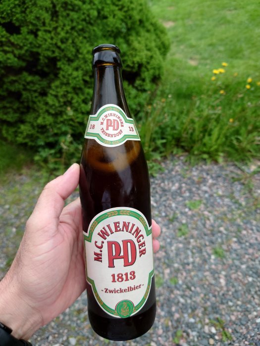 Hand håller en flaska Zwickelbier med trädgård i bakgrunden.