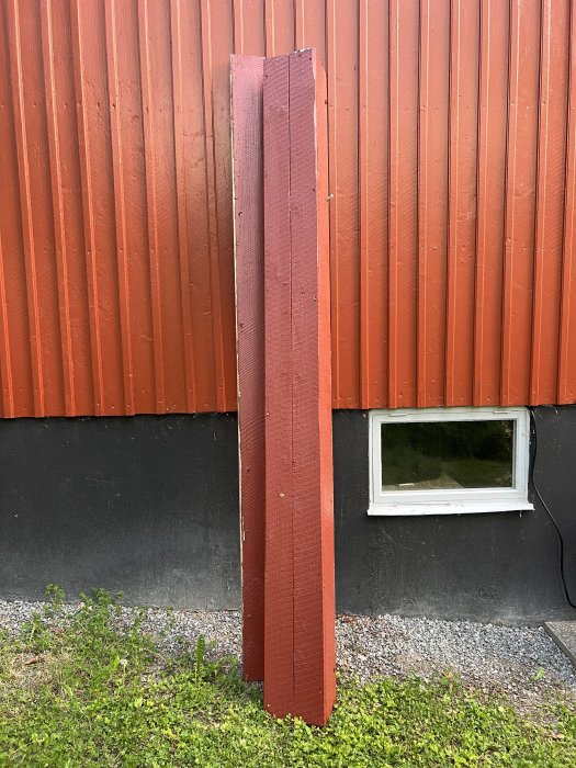 Rödmålad träplanka står lutad mot en röd trävägg vid ett hus med ett litet fönster.