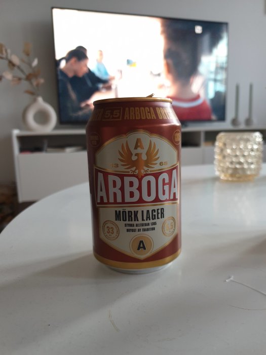 En burk Arboga Mörk Lager framför en suddig TV som visar en fotbollsmatch.