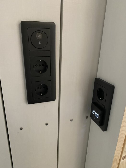 Svart modern väggkontakt och strömuttag monterade på ljusgrå panelvägg, med en digital temperaturdisplay.