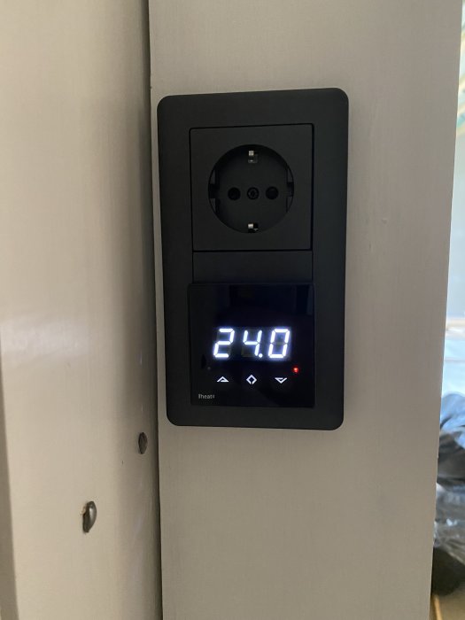 Digital termostat vid 24 grader med touchknappar monterad på vägg ovanför strömuttag.