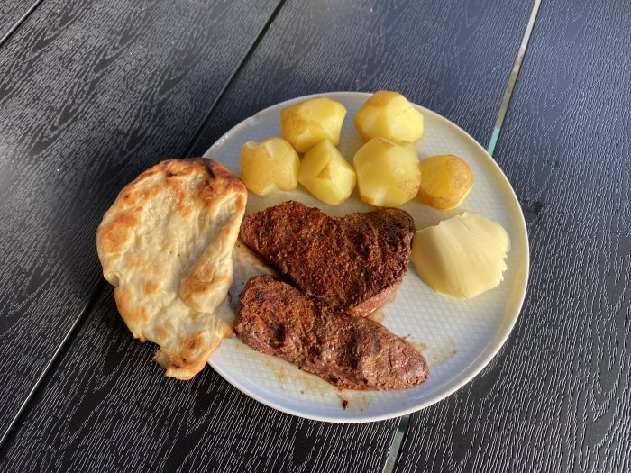 Tallrik med grillade biffar, kokt potatis, smält smör och ett naanbröd på ett bord.