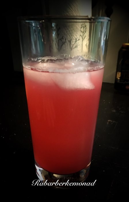 Ett glas fylld med hemgjord rosa rabarberlemonad med isbitar.