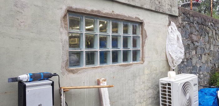 Fasadrenovering pågår med putsade ytor runt ett glasblocksfönster, verktyg och värmepump i förgrunden.