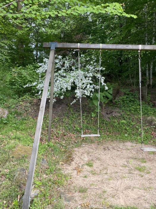 Gunga med metallkedjor och trästolpar framför en jordhög under ett träd med vita blommor.