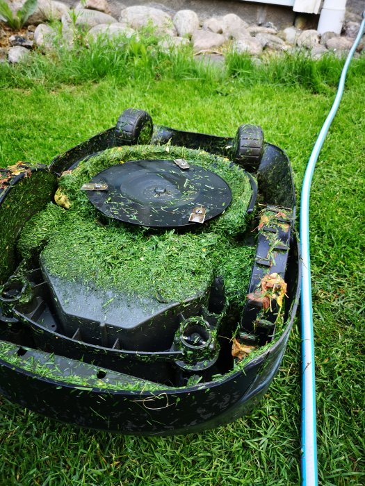 Gräsklippare täckt av gräsrester och våta löv efter klippning i regn, står på blöt gräsmatta.