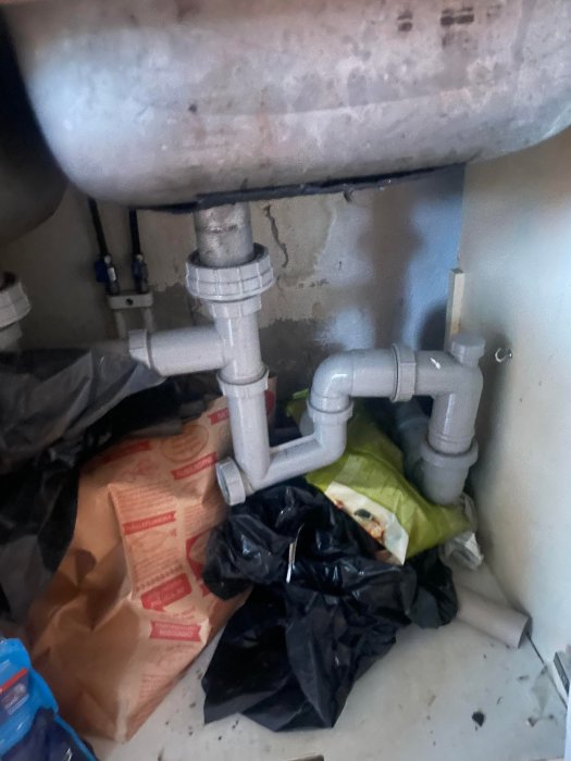 Rörsystem under en diskbänk med komplex anslutning, intill en betongvägg och oorganiserade rengöringsmedel.