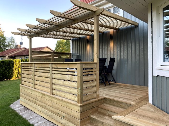 Robust pergola i trä med takreglar belägen vid ett hus, omgiven av en trädgård och utrustad med en uteplats och sittmöbler.