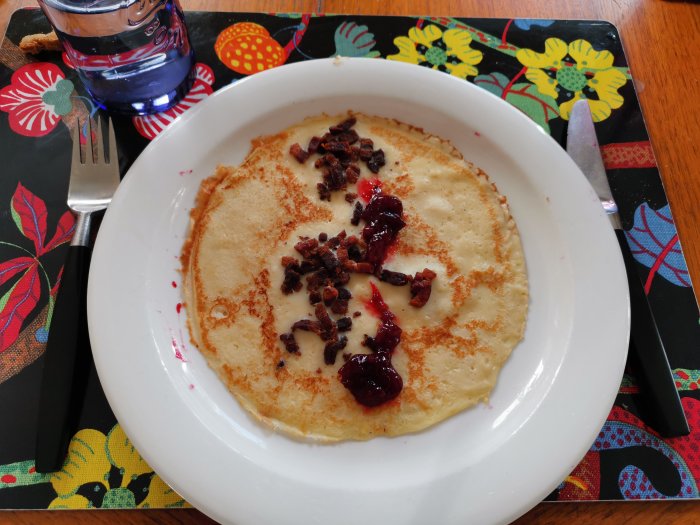 Tunn pannkaka på tallrik med knaperstekt fläsk och röd sylt, vatten och bestick vid sidan.