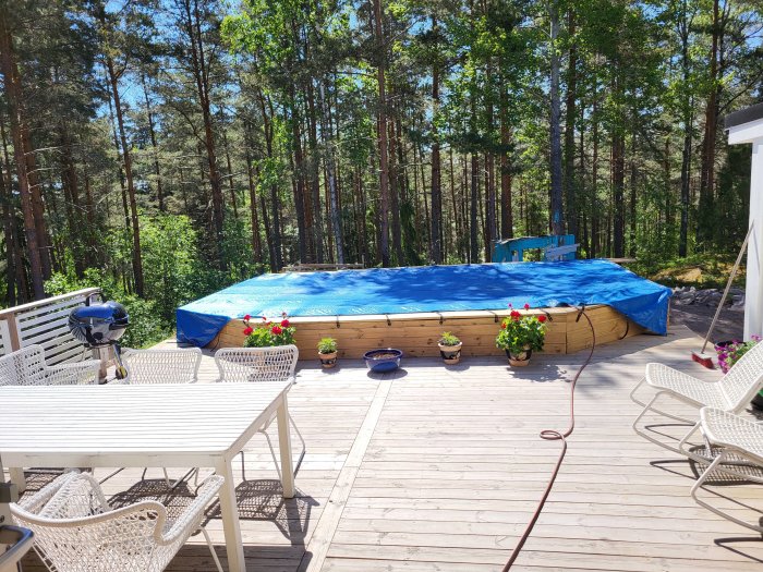 Uppbyggd trädäck med överdragtäckt pool, blomkrukor och utemöbler på solig dag med skog i bakgrunden.