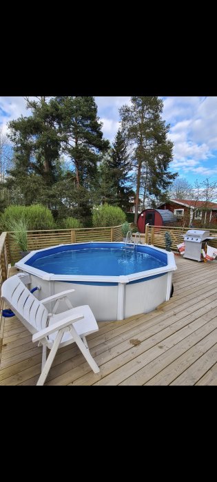 Rund ovanmarkspool på trädäck med vit solstol, grill och gröna träd i bakgrunden.