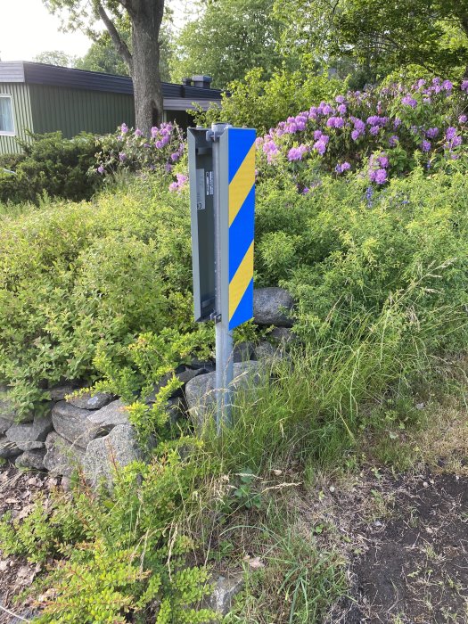 Stolpe på cirka 150 cm med dubbla blå och gula vägskyltar i en trädgård.