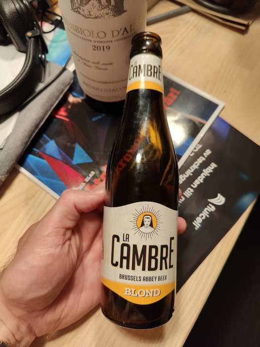 Hand håller en ölflaska med etiketten "La Cambre Brussels Abbey Beer Blond" framför en vinflaska och böcker.